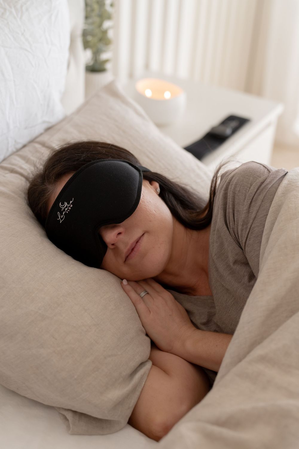 Woman sleeping peacefully in bed wearing a black sleep eye mask, resting on neutral linen pillows with candle in background.