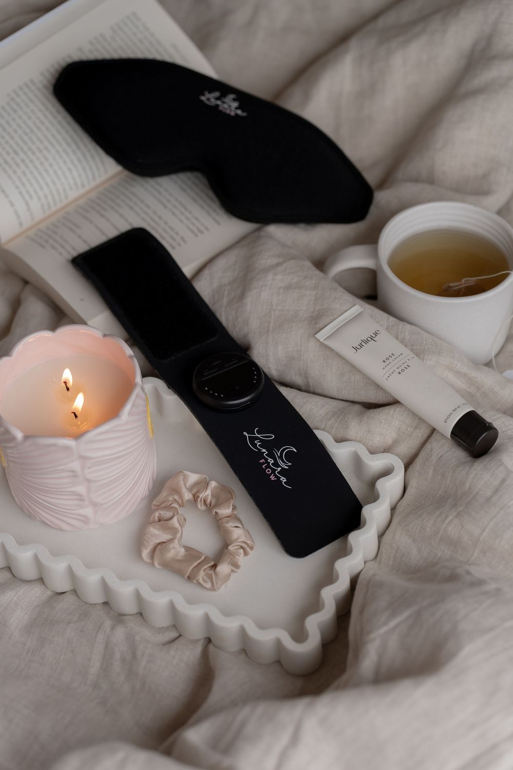 Cozy bedtime flatlay with sleep mask, hair wrap, candle, scrunchie, tea, and skincare on a ruffled tray.