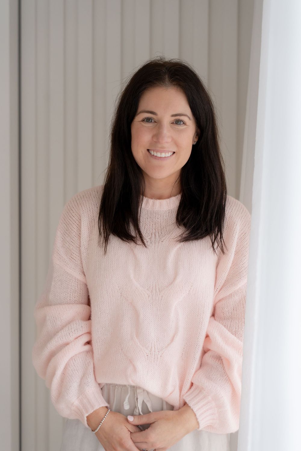 Smiling woman with dark hair wearing a soft pink knit sweater, standing against a light neutral background.