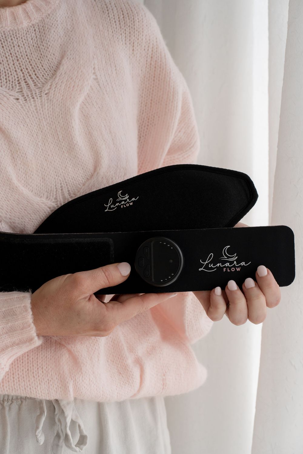 Woman in pink knit sweater holding a black Lunara Flow device and case with both hands.