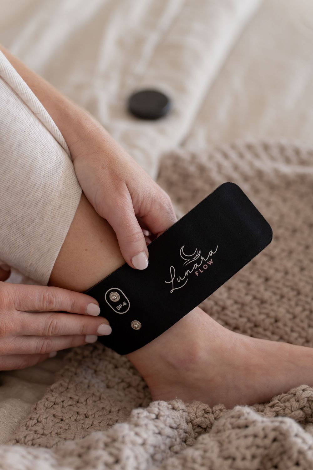 Person holding a black Luna Flow tool massager device while sitting on a cozy knit blanket.