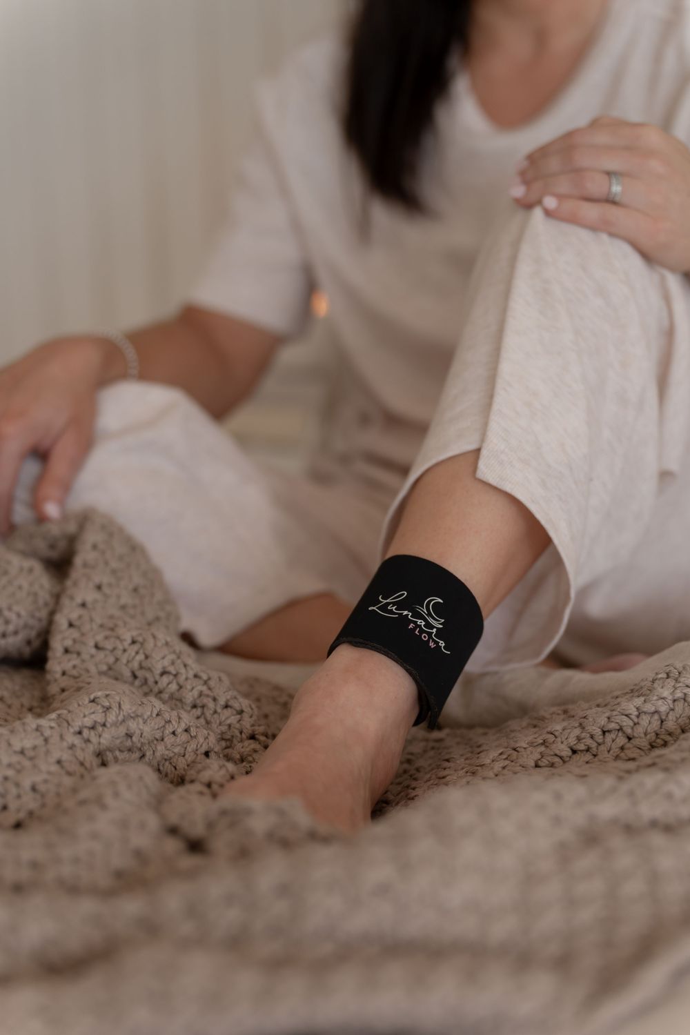 Woman sitting on bed wearing a black ankle band/bracelet with logo, dressed in white linen outfit with cozy knit blanket.