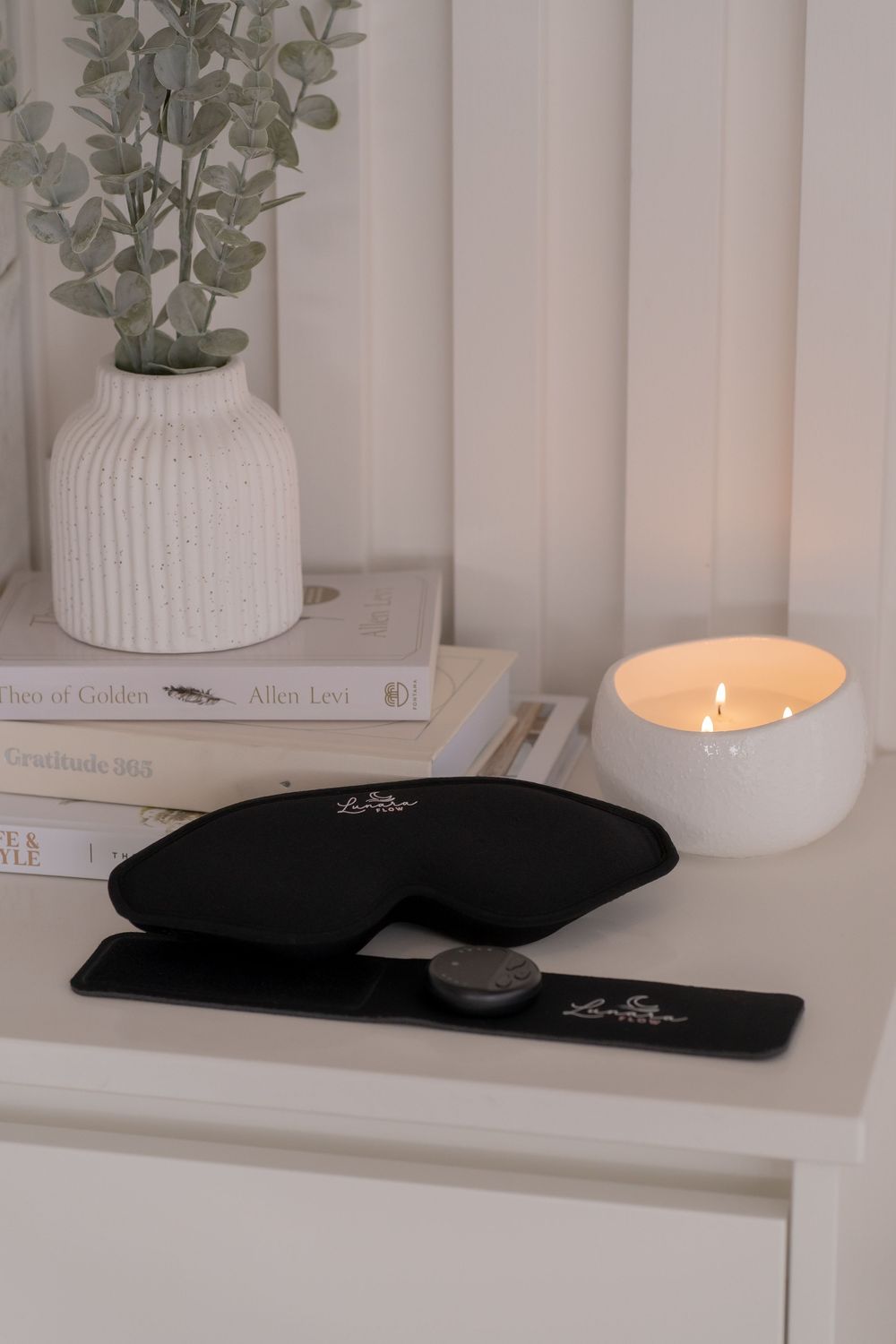 Black silk sleep mask and eye pillow on white nightstand with lit candle, books, and eucalyptus vase.
