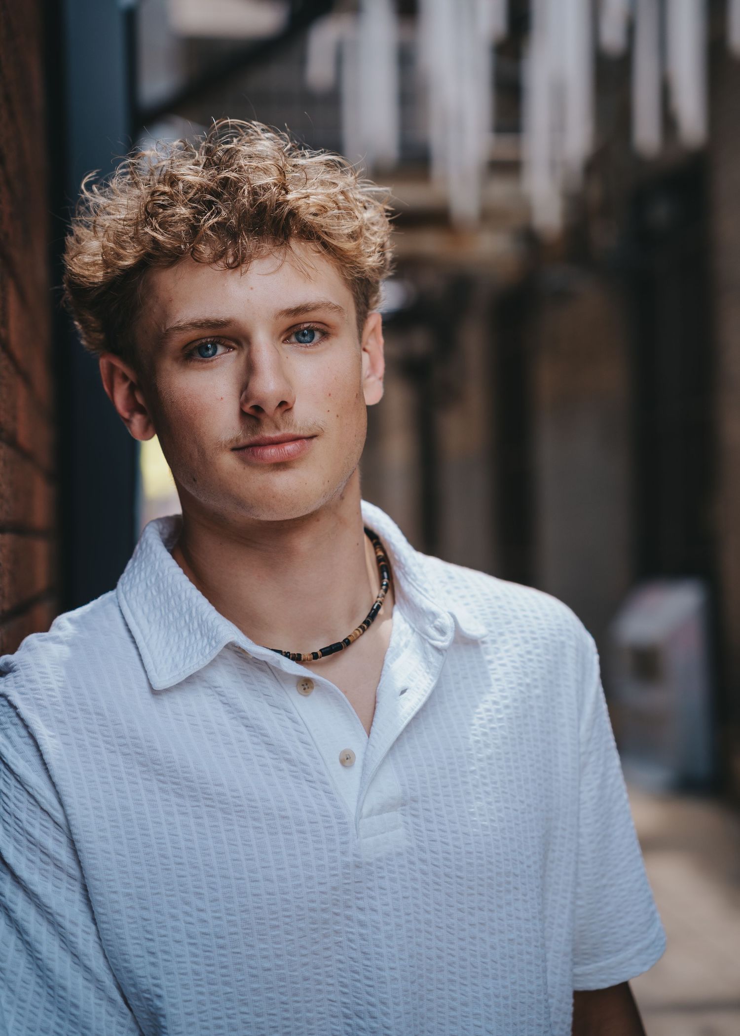 Young man with curly blonde hair wearing a white polo and beaded necklace poses in an urban alley.