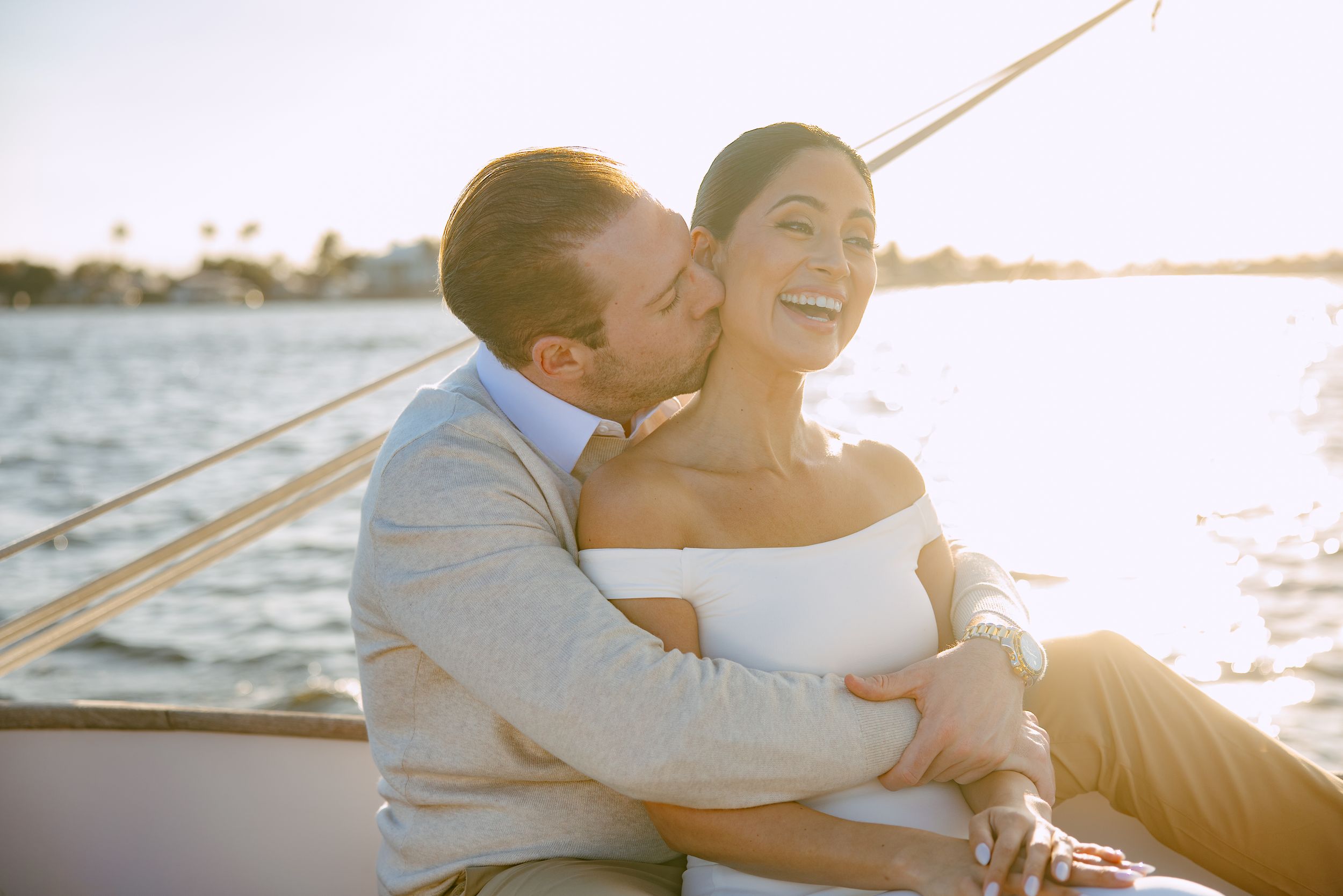 Man kissing smiling woman on a sailboat at sunset, both dressed in elegant attire.