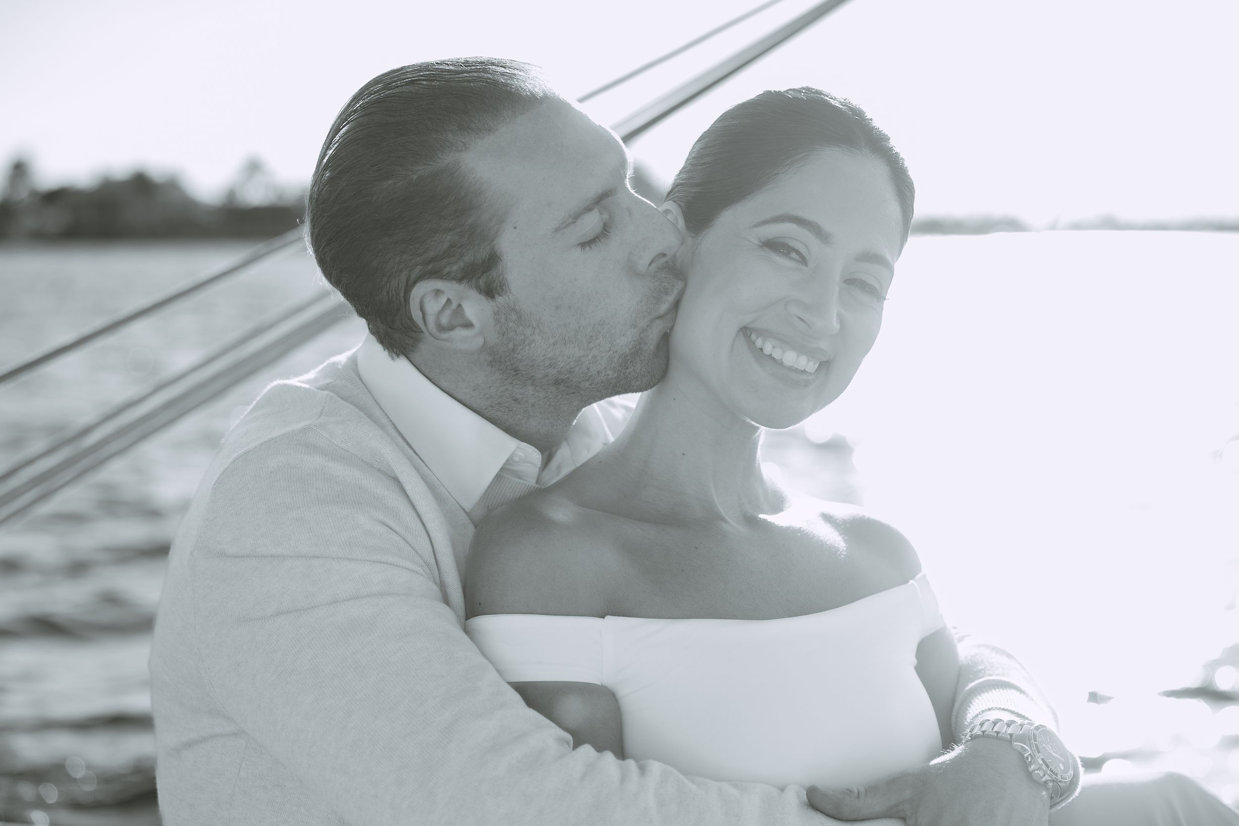 Black and white photo of a couple sharing a romantic kiss on a sailboat on the water.