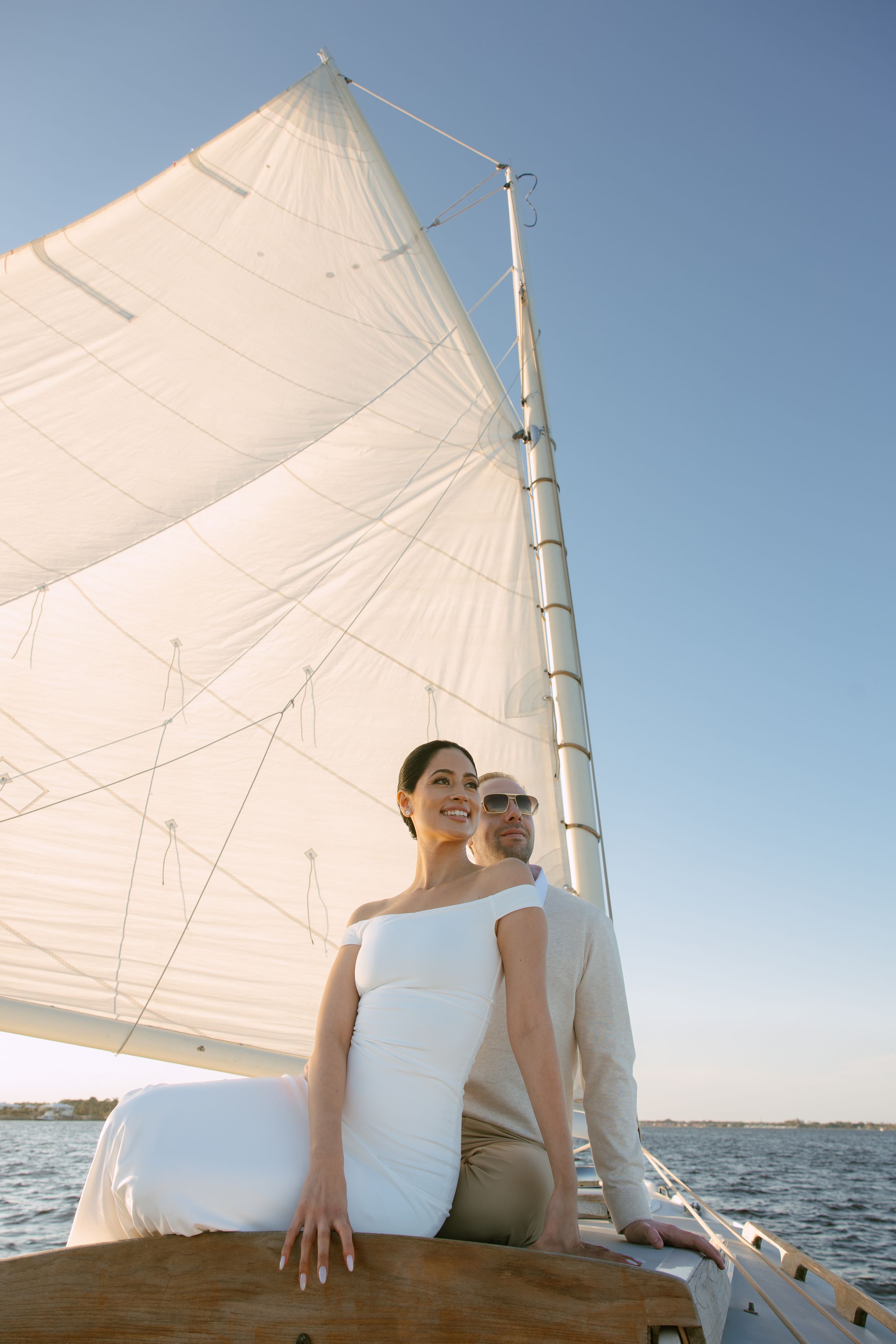 Bride and groom posing on the bow of a sailboat with white sails against a clear blue sky.