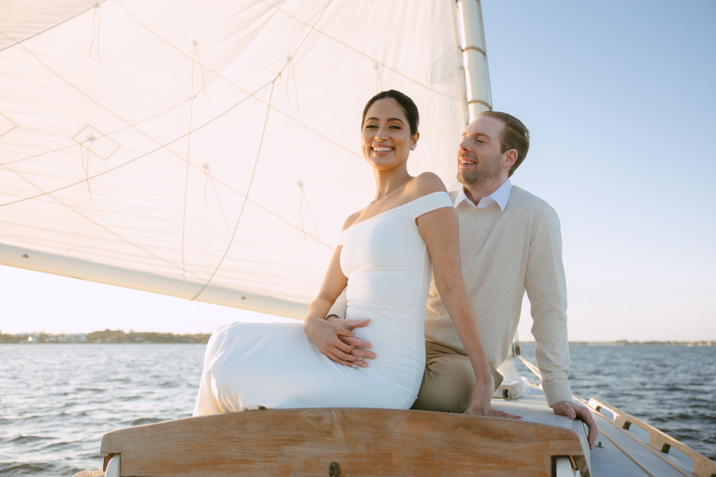 Smiling bride in white dress sitting with groom on sailboat deck at golden hour.