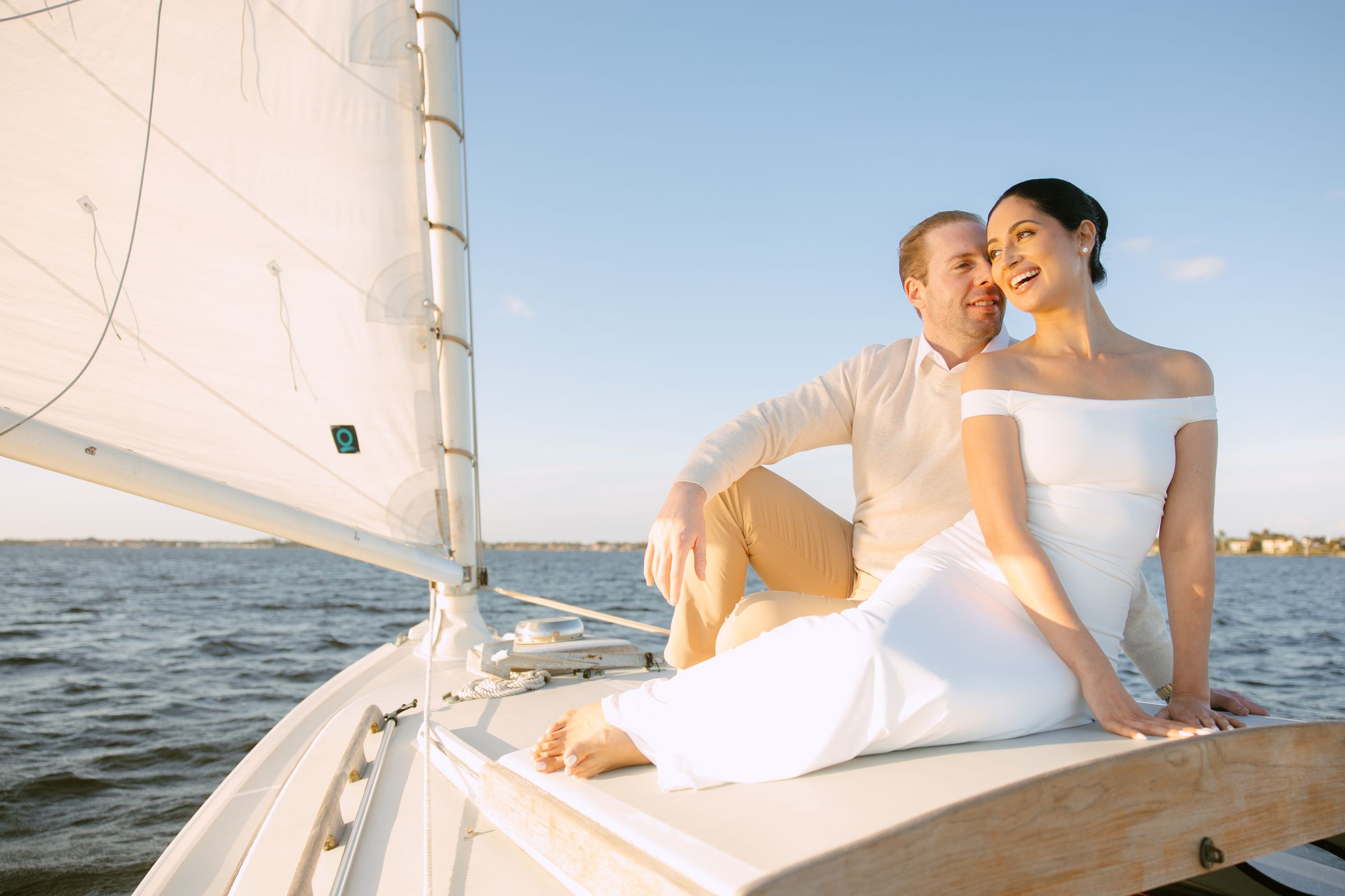 Couple in wedding attire relaxing together on sailboat bow with water and sails behind them.