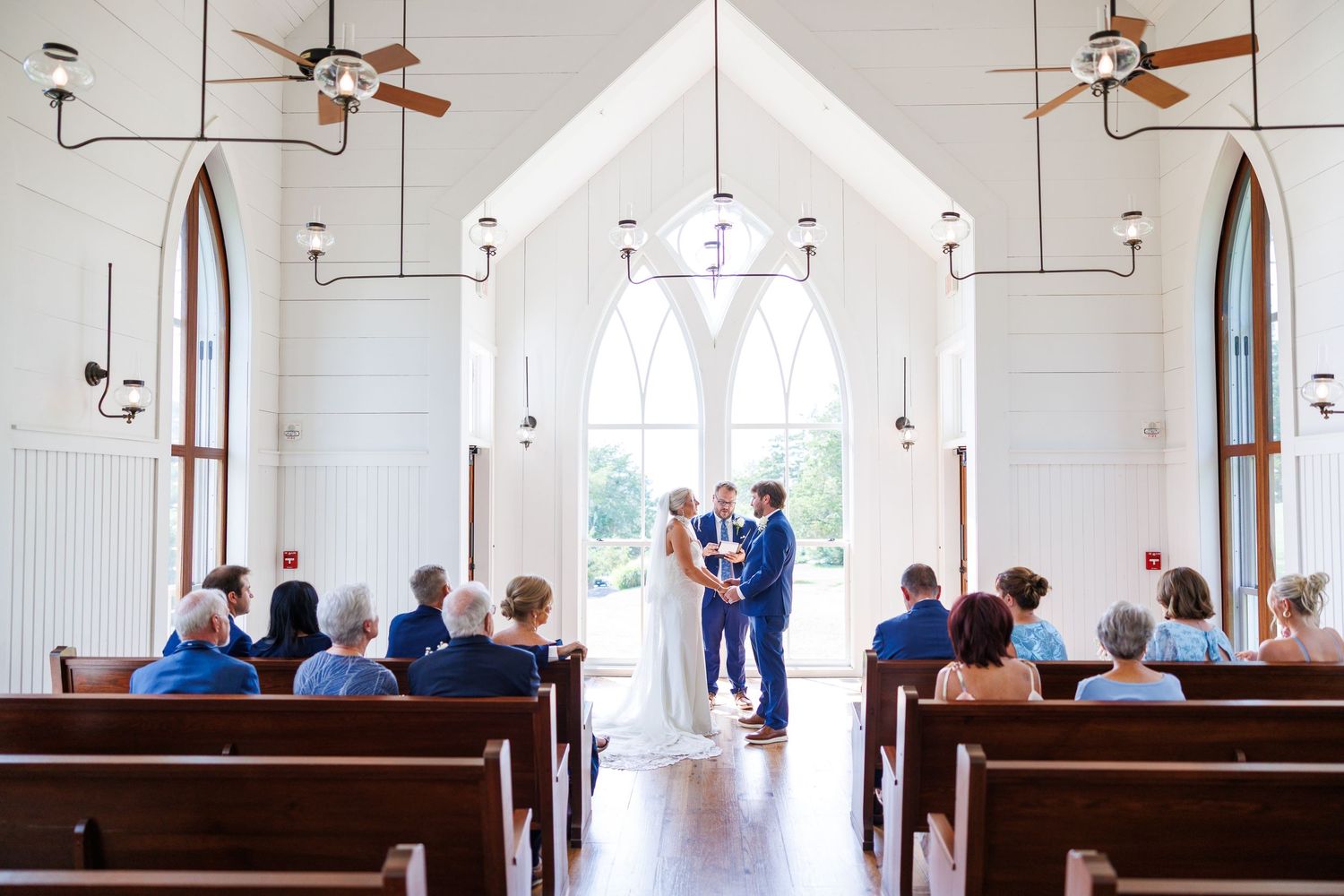 Couple exchanges vows at elegant white chapel wedding ceremony with arched windows and wooden pews filled with guests.
