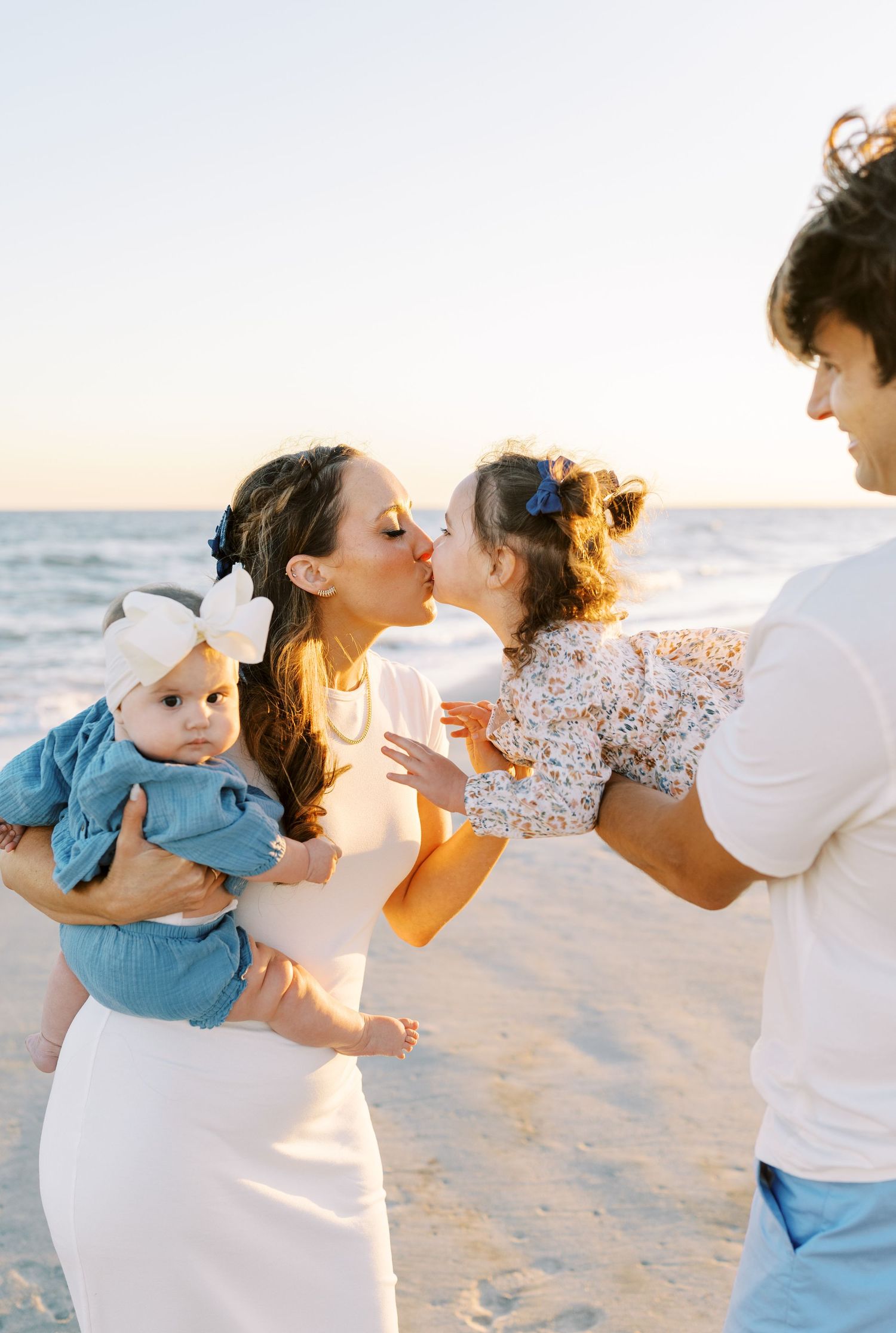 Family beach photo at sunset with mom kissing toddler held by dad while cradling baby in white dress.
