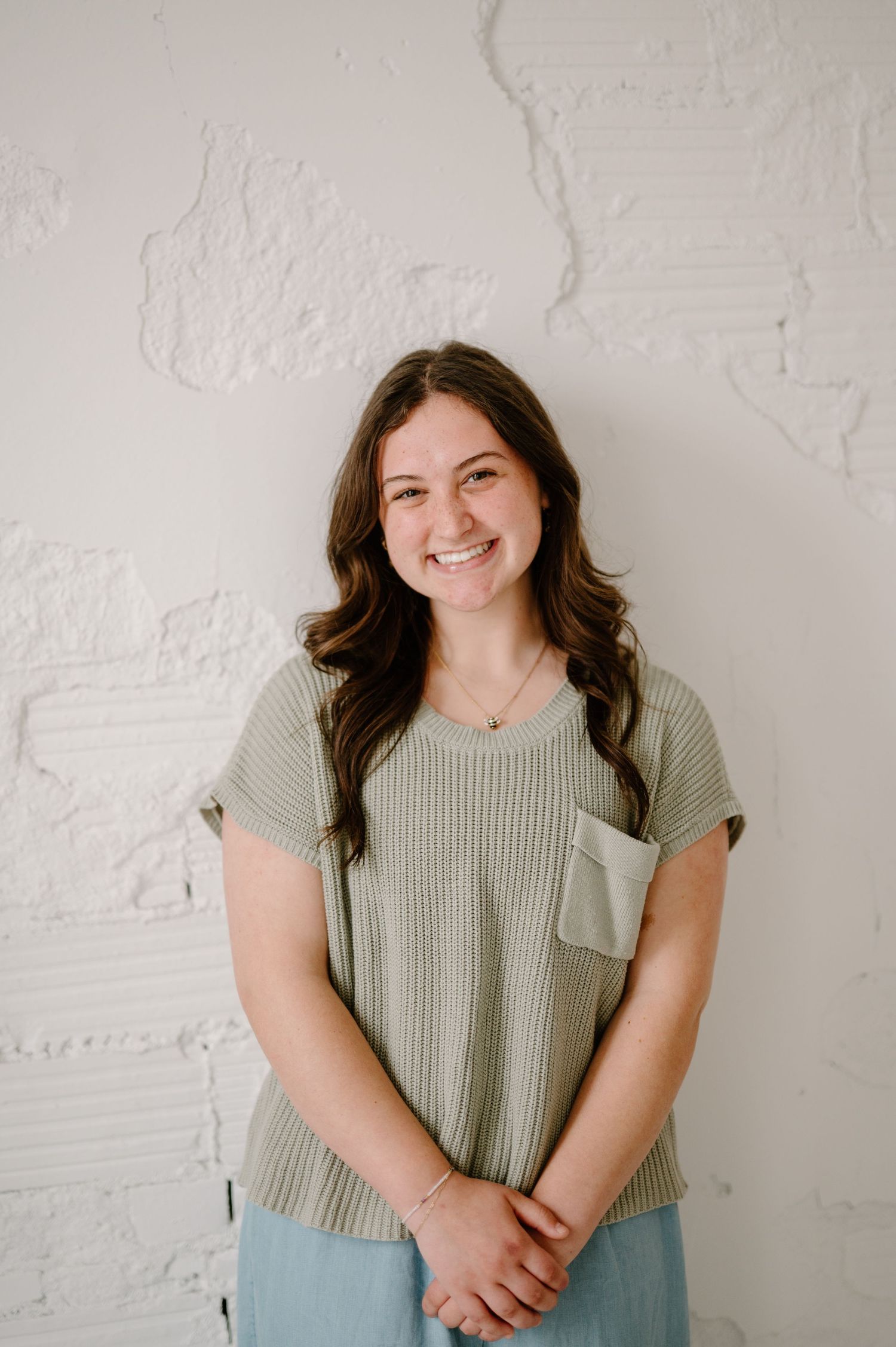 Young woman with long brown hair smiling, wearing a sage green ribbed top and blue pants, standing against a white textured wall.