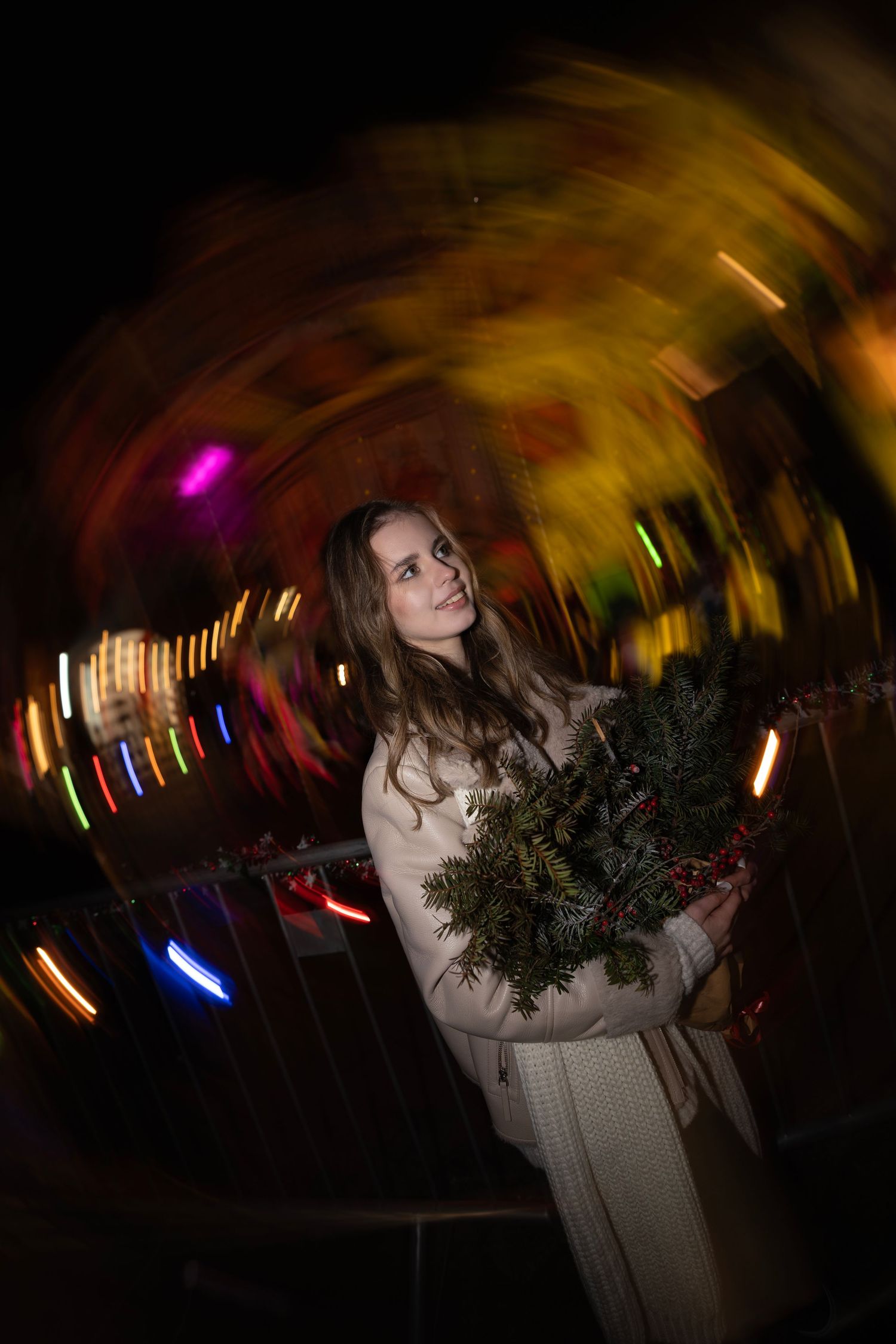 Woman in white coat holds holiday pine wreath surrounded by swirling colorful bokeh night lights.