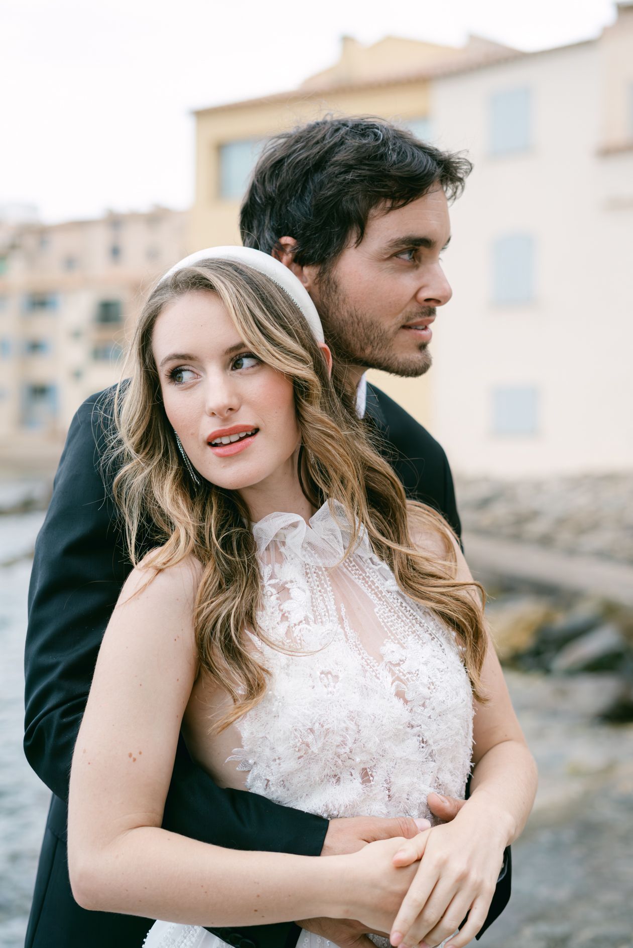 A bride and groom embrace outdoors near a waterfront, she wears a lace dress and headband, he wears a dark suit.