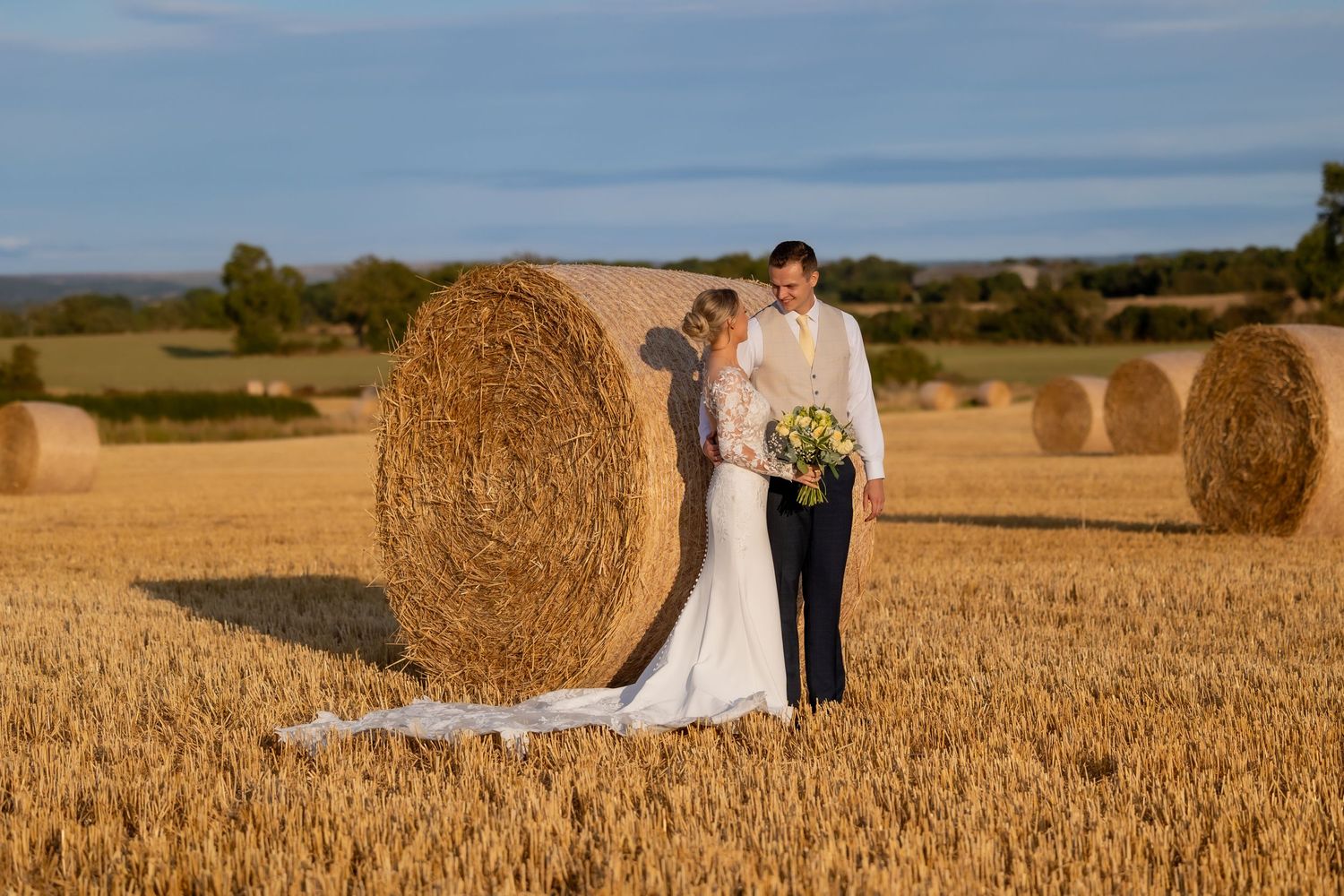 Bride and groom posing beside hay bales in a golden wheat field on their wedding day. Casual Captures UK