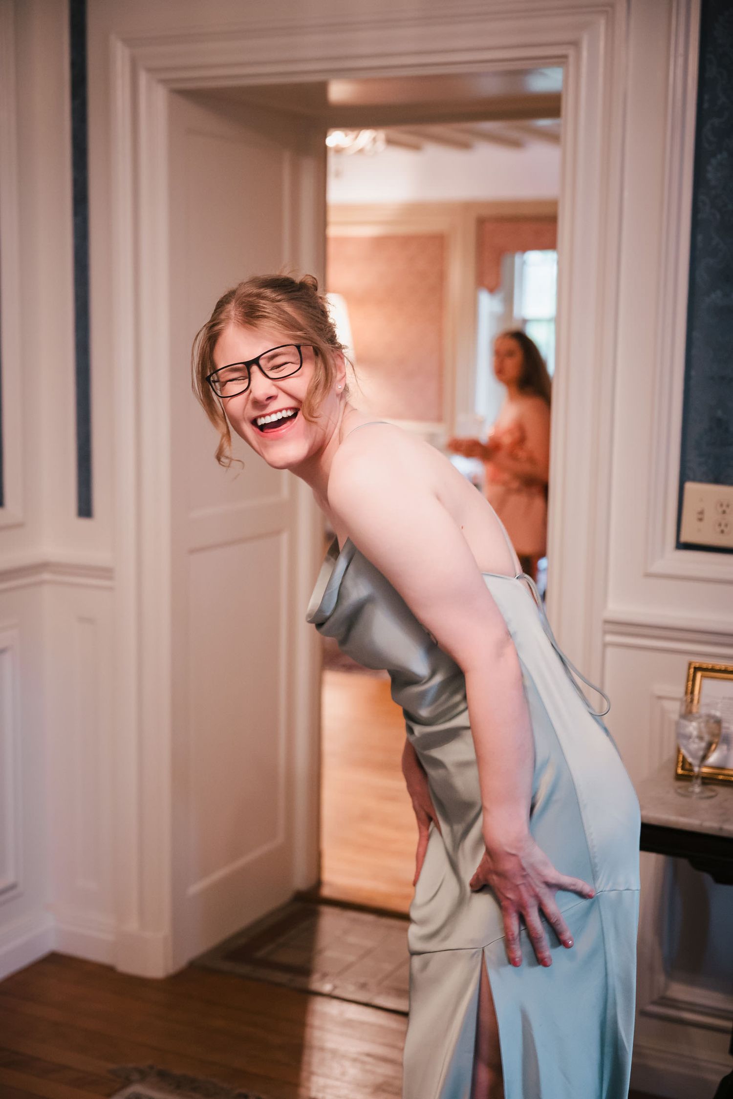 Bridesmaid laughing candidly in a sage satin dress during wedding day preparations, with the bride visible behind her.