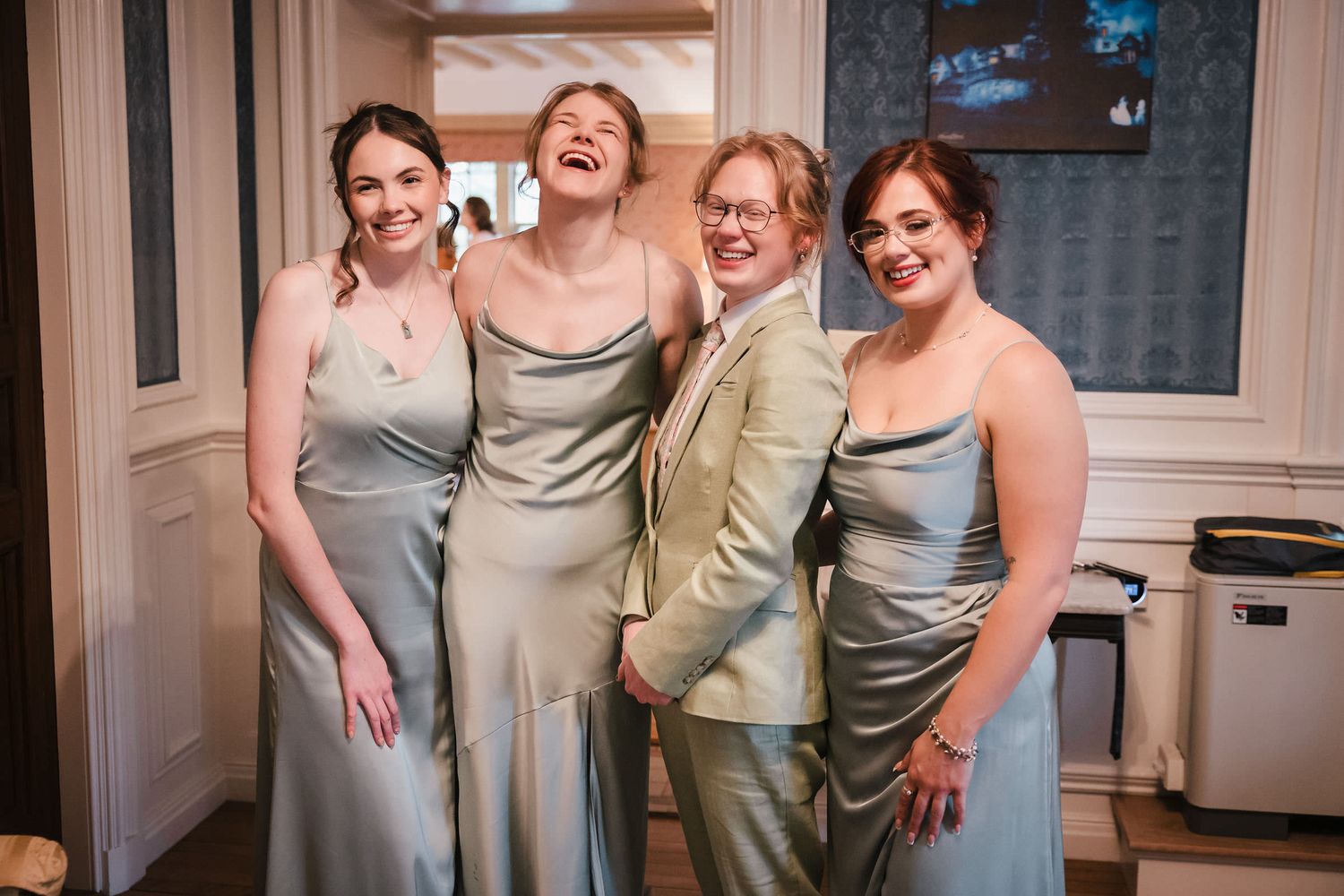 Four laughing bridesmaids, one in a suit, standing together during wedding day preparations at Searles Castle