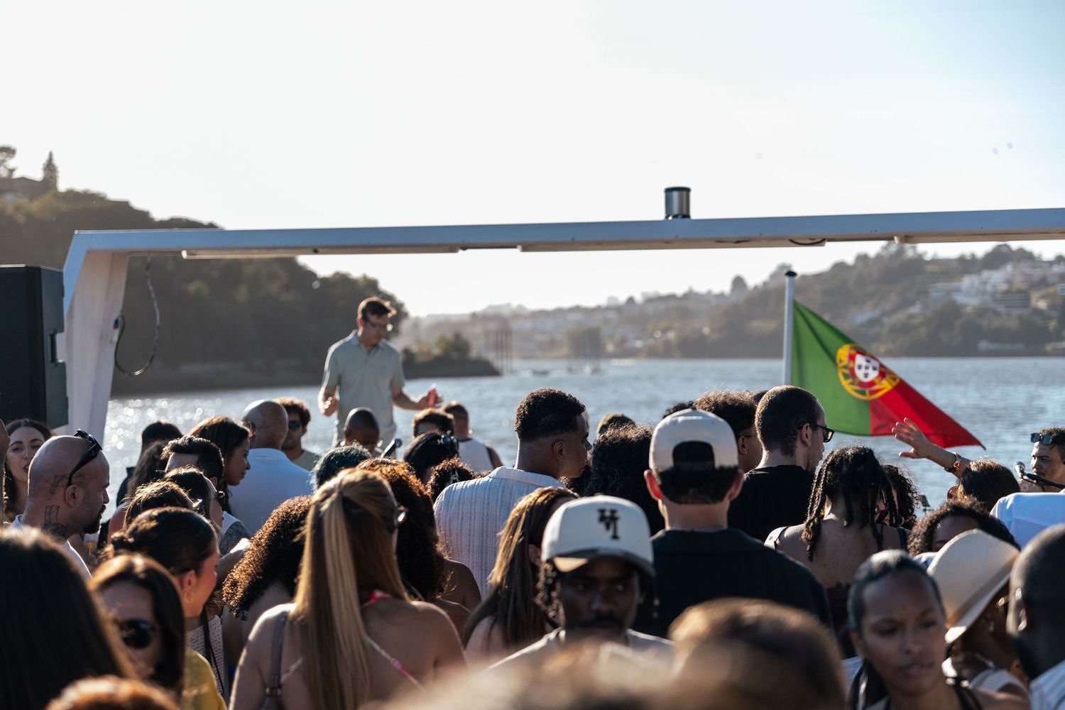 Crowd enjoying an outdoor party near the water with a Portuguese flag waving in the sunlight.