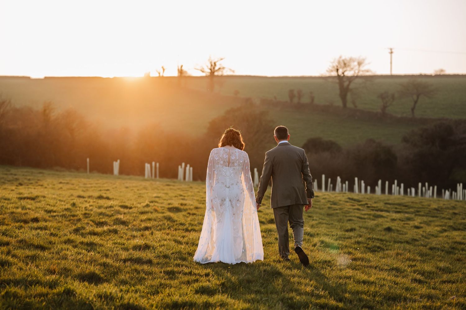 Couple holding hands running through golden wildflower field at sunset in wedding attire at The Cornish Place