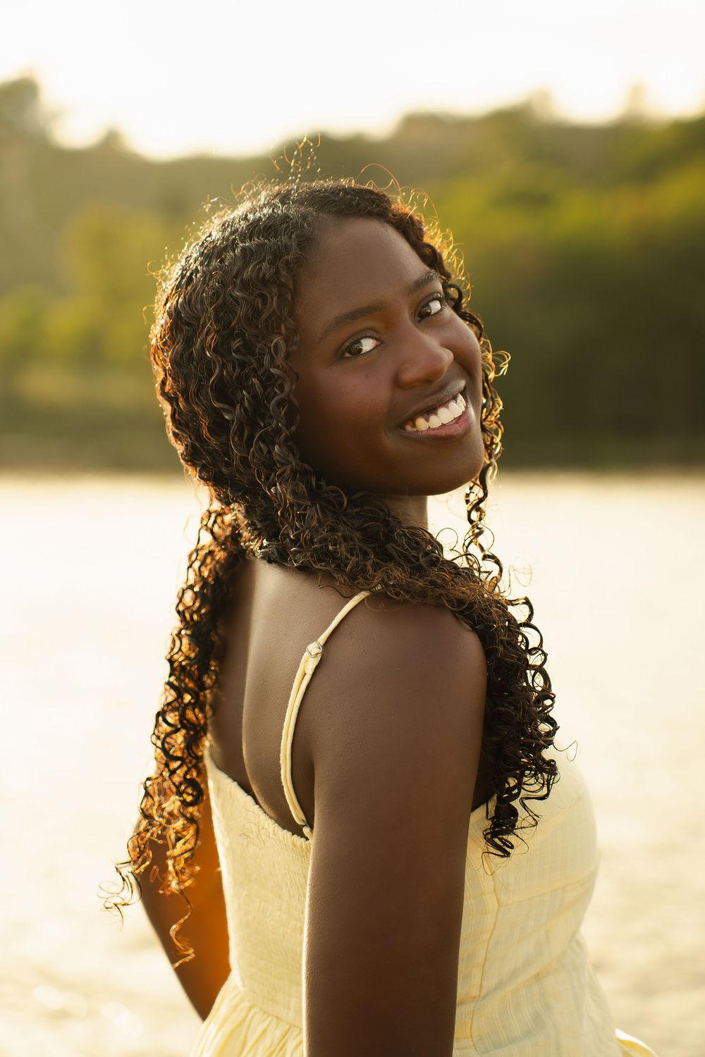 Smiling young Black woman with long curly hair wearing a yellow dress outdoors near water at golden hour.
