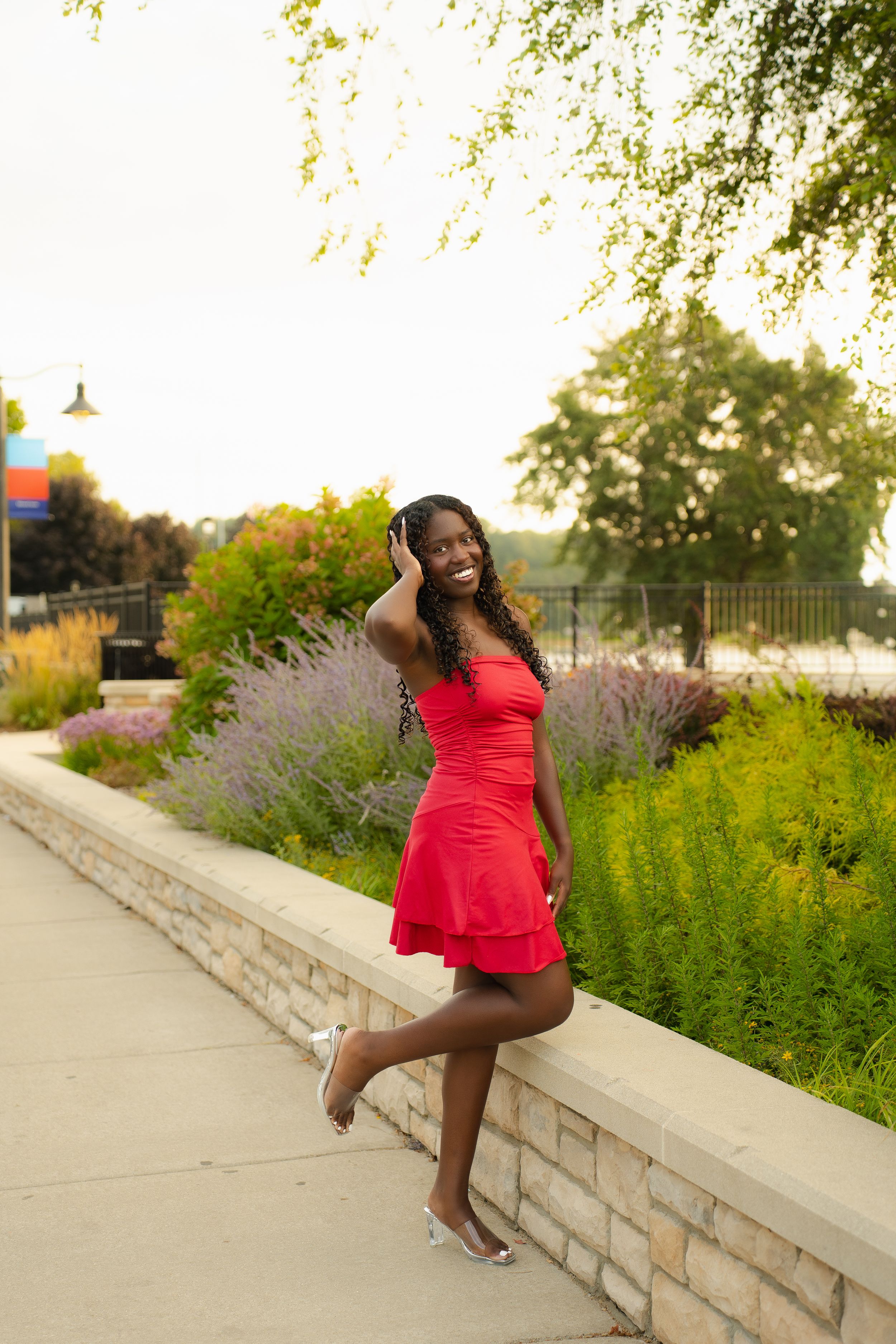 Young woman in red dress posing outdoors by a stone wall with colorful garden background.