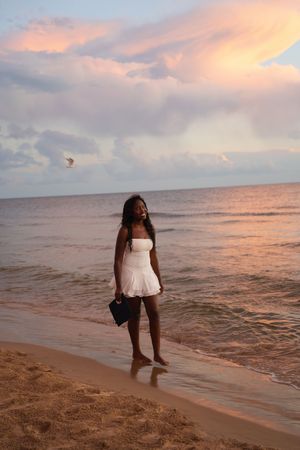 A young woman in a white dress stands barefoot on a sandy beach at sunset with pink clouds over the ocean.