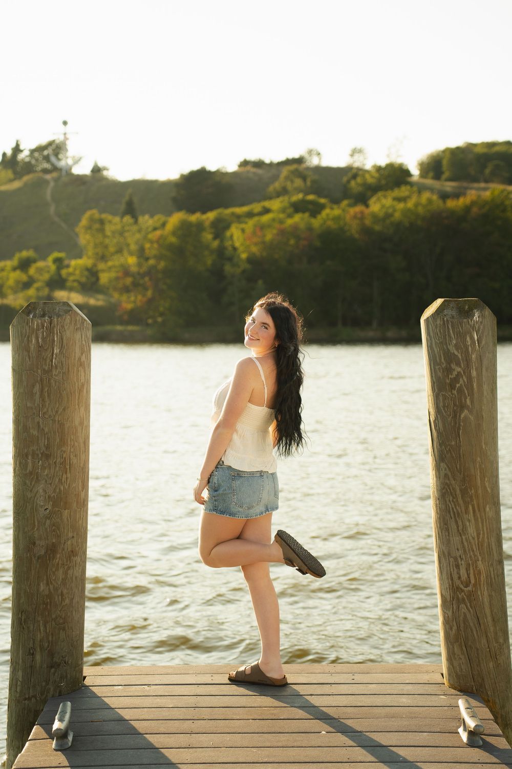 Young woman in denim shorts and white tank top posing on a wooden dock by a lake, smiling over her shoulder.