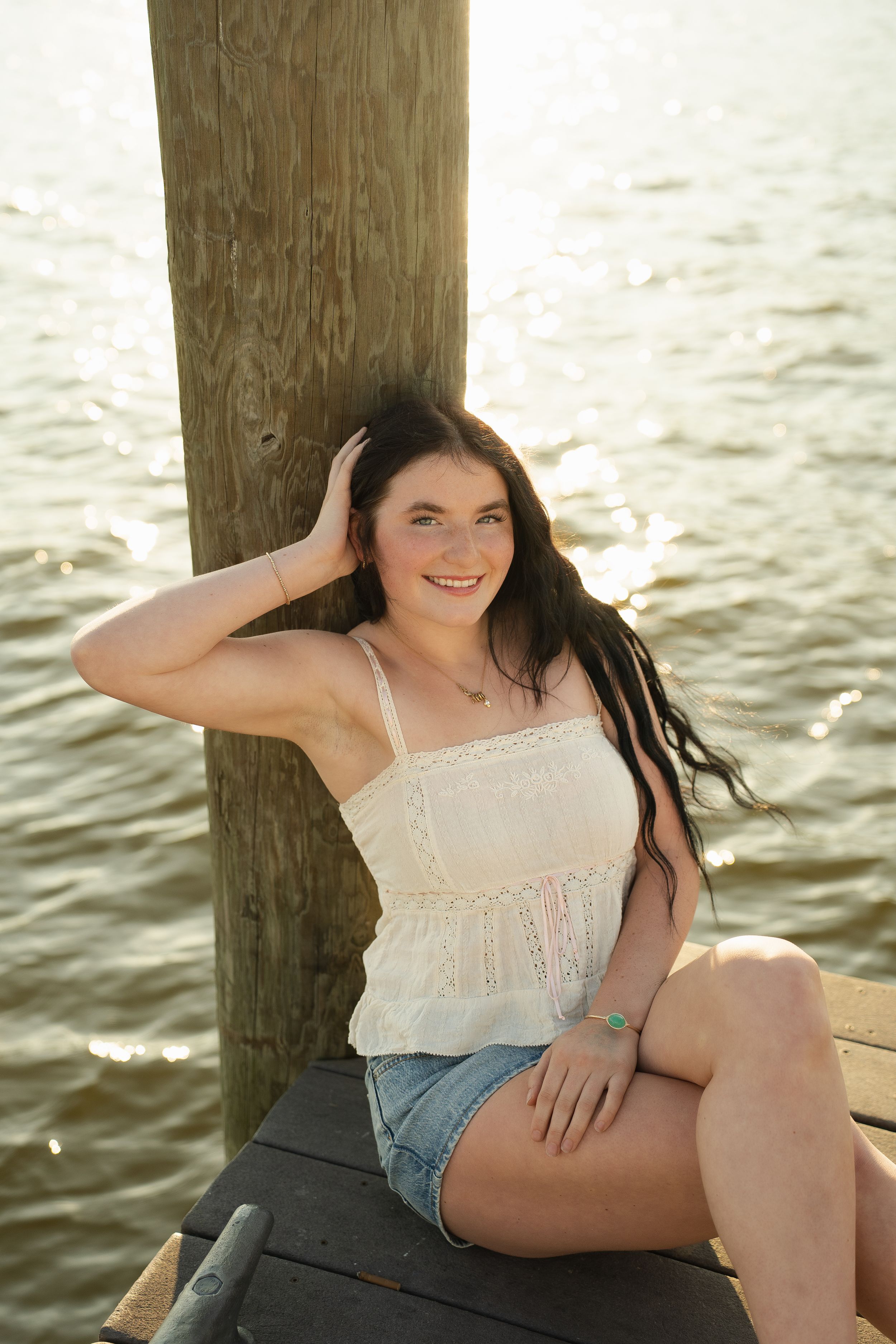 Young woman in white top and denim shorts sitting on a wooden dock by the water, smiling in golden light.