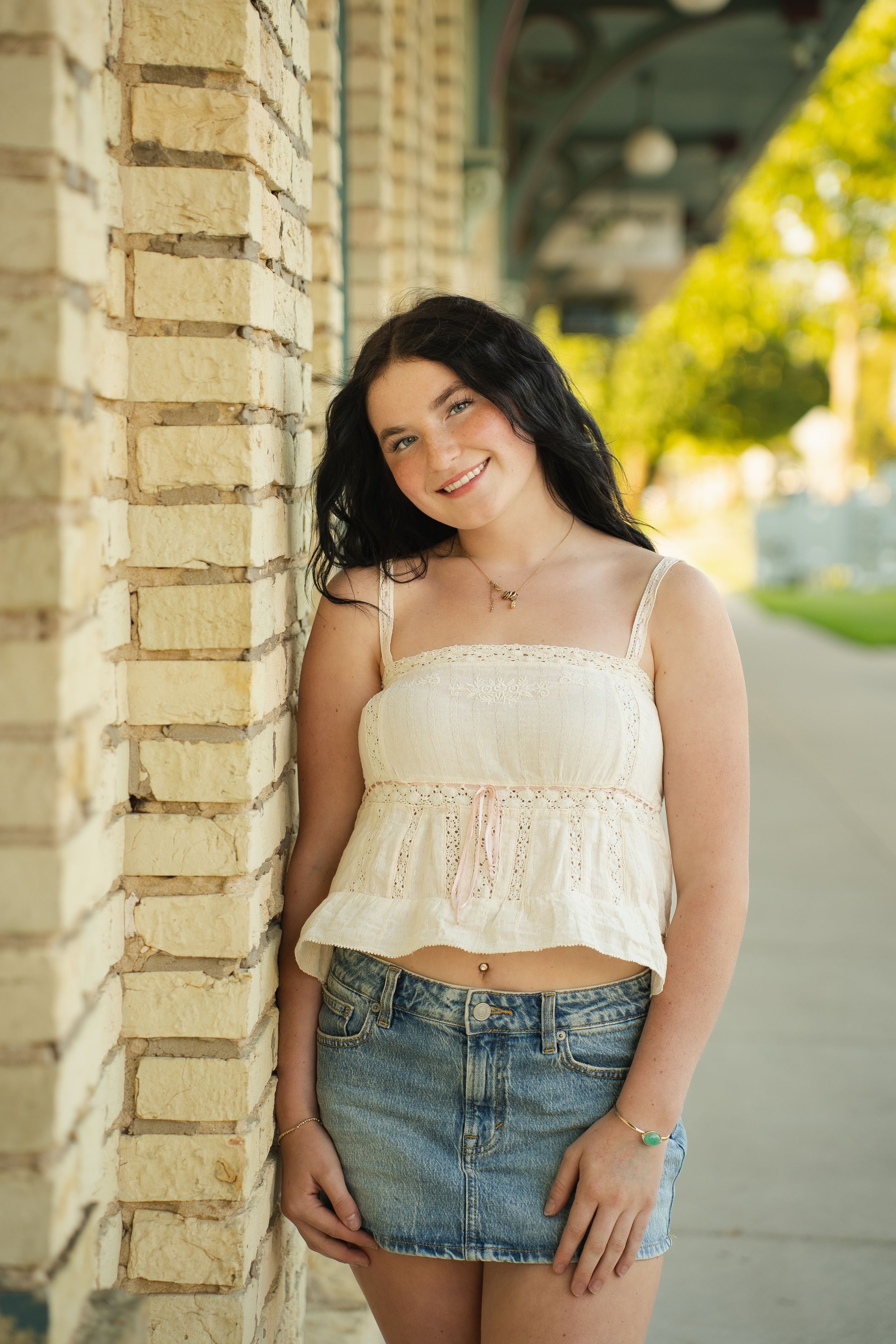Young woman in white crop top and denim skirt smiling against brick wall outdoors.