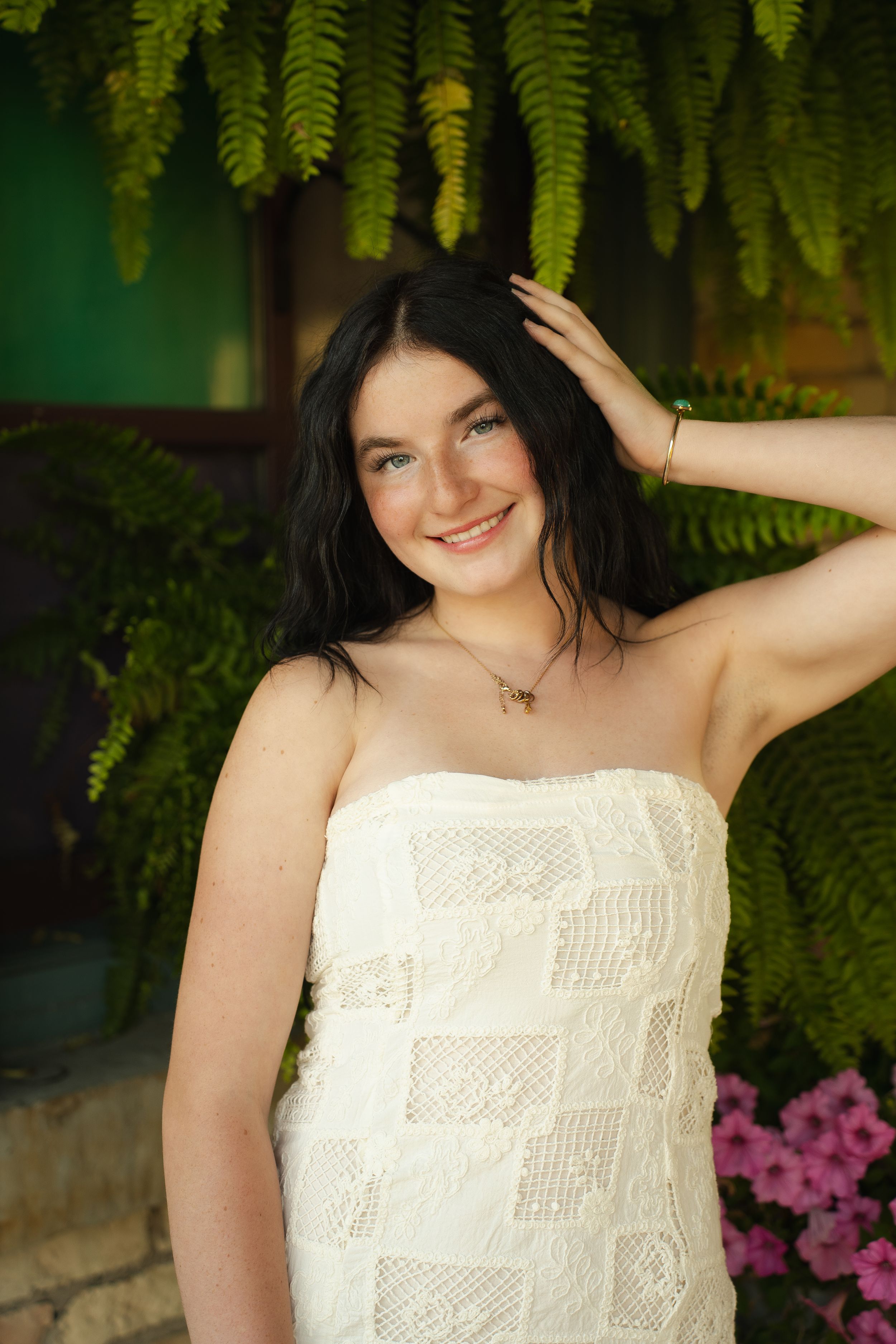 Young woman with dark hair smiling in white strapless dress, posing among lush green ferns and pink flowers.