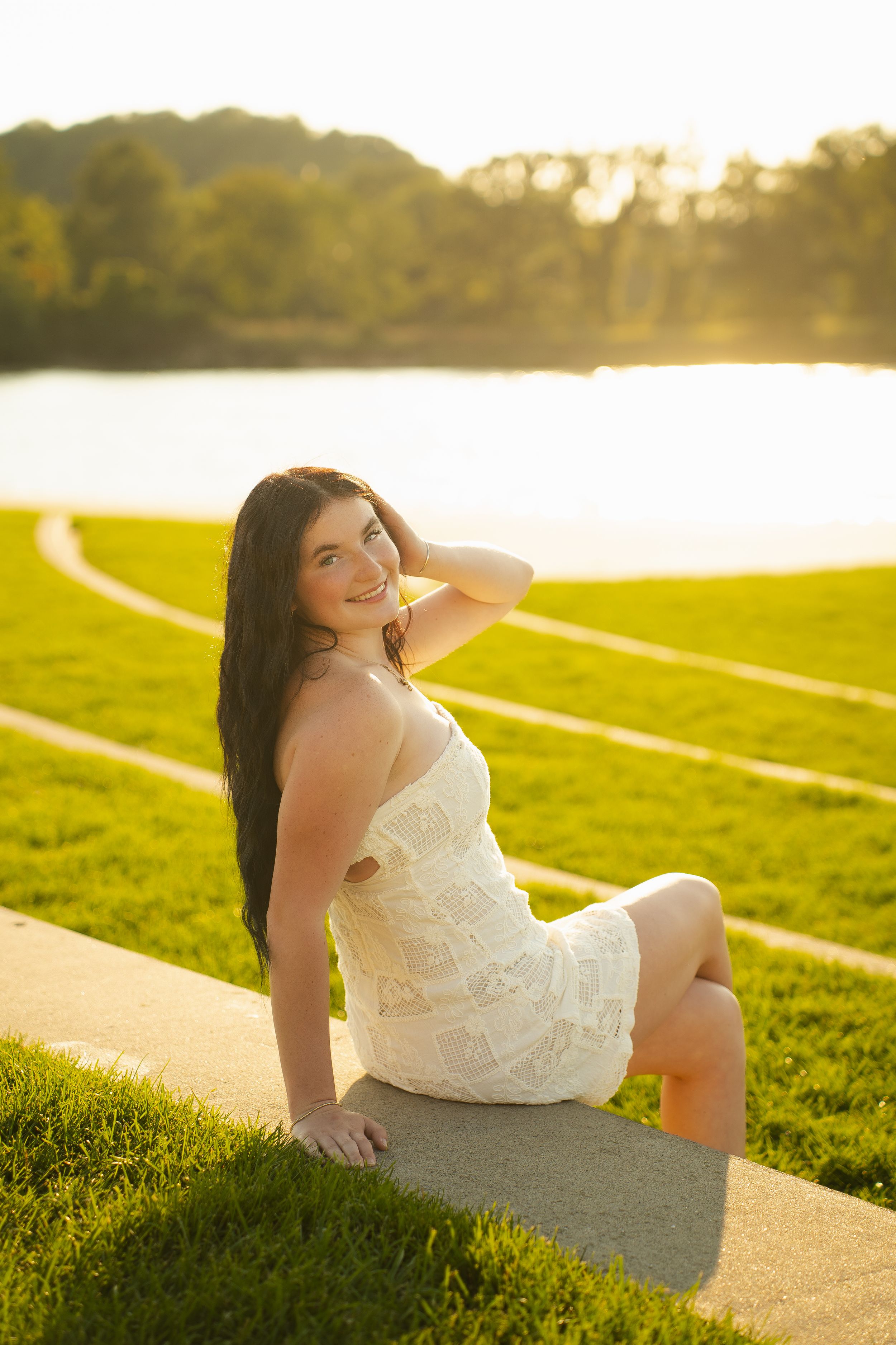 Young woman in white lace dress sitting on a path in a sunlit park with a lake and green trees behind her.