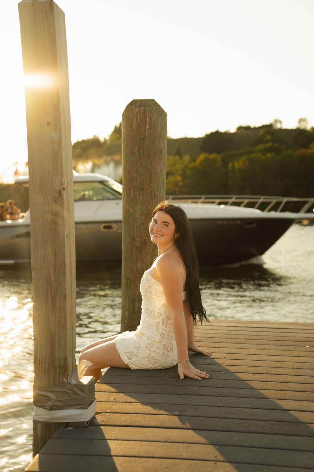 Young woman in white dress sitting on a wooden dock at golden hour with a boat on the water behind her.