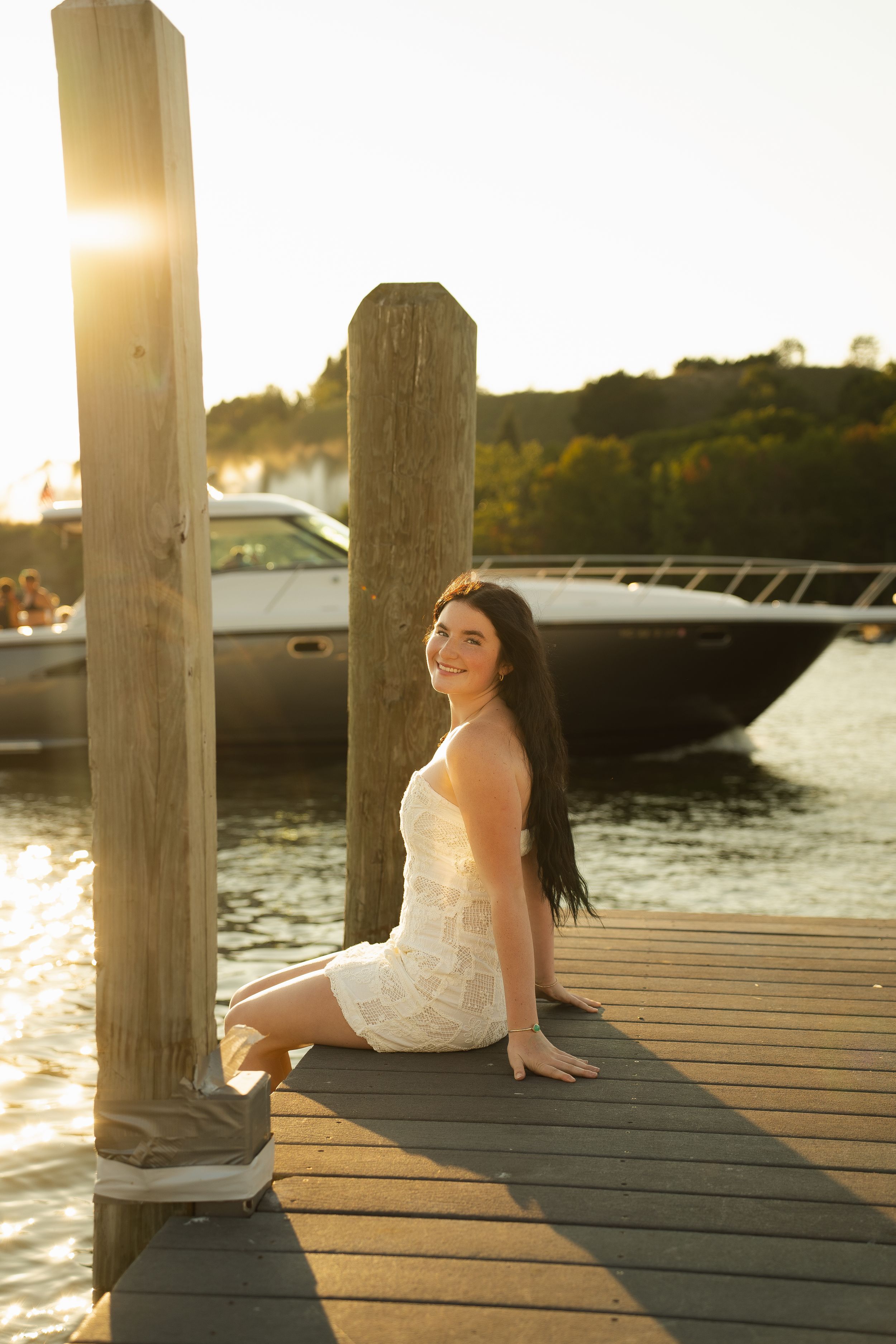 Young woman in white dress sitting on a wooden dock at golden hour with a boat on the water behind her.