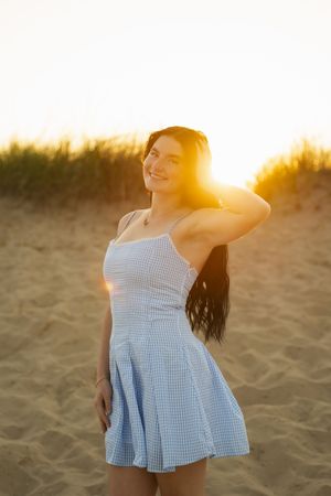 Young woman in blue gingham sundress poses on sandy beach at golden sunset with warm lens flare.