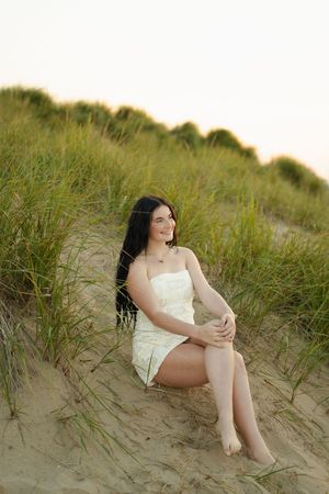 Young woman in white dress sitting on sandy beach dunes with green grass, smiling at sunset.