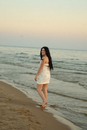 Young woman in white dress walking barefoot on sandy beach at sunset with ocean waves behind her.