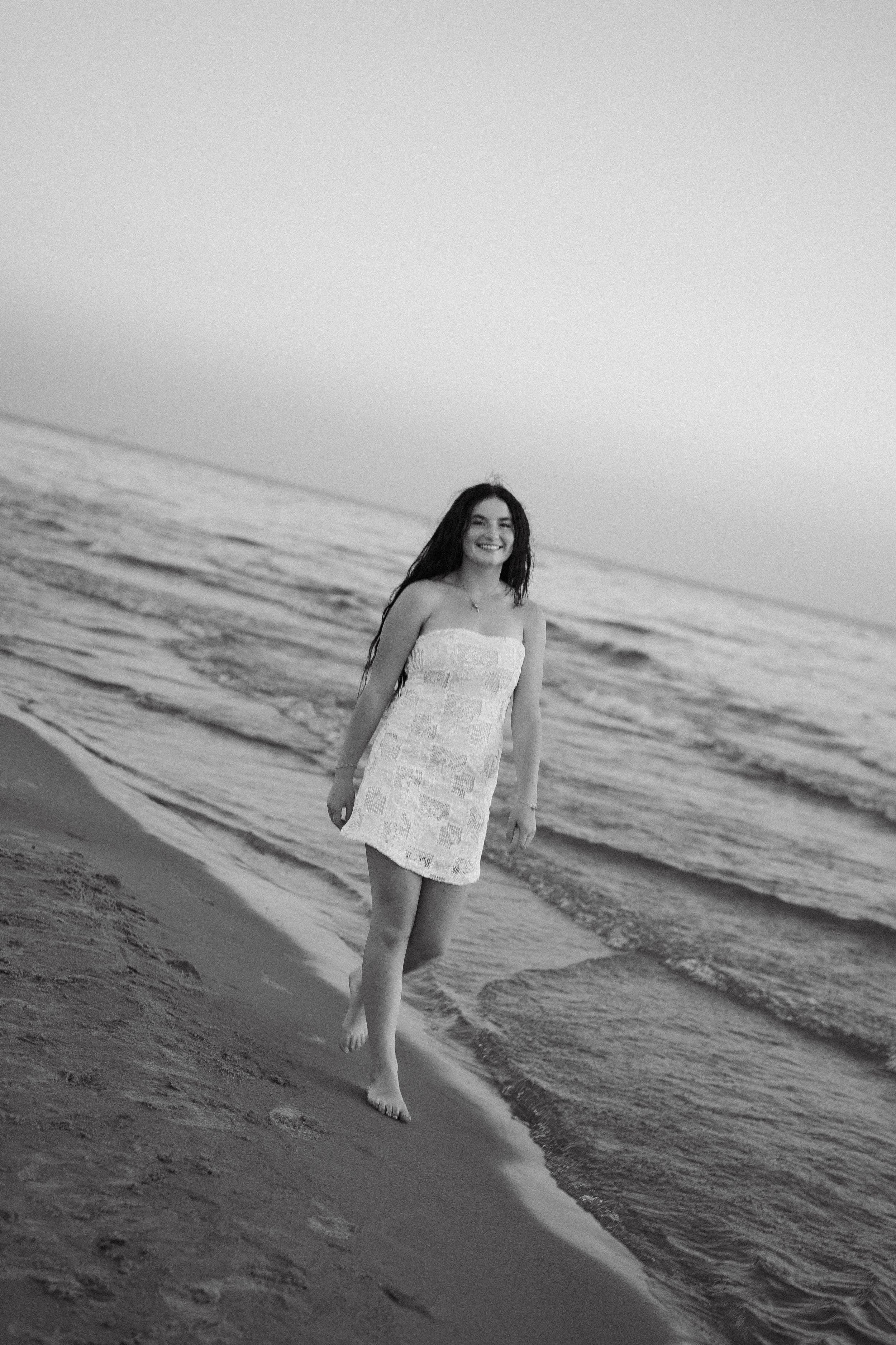 Black and white photo of a smiling young woman in a white dress walking barefoot along a sandy beach shoreline.