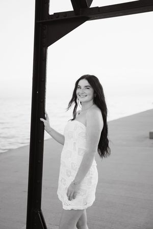 Young woman in white strapless dress smiling near metal structure by the ocean in black and white photo.