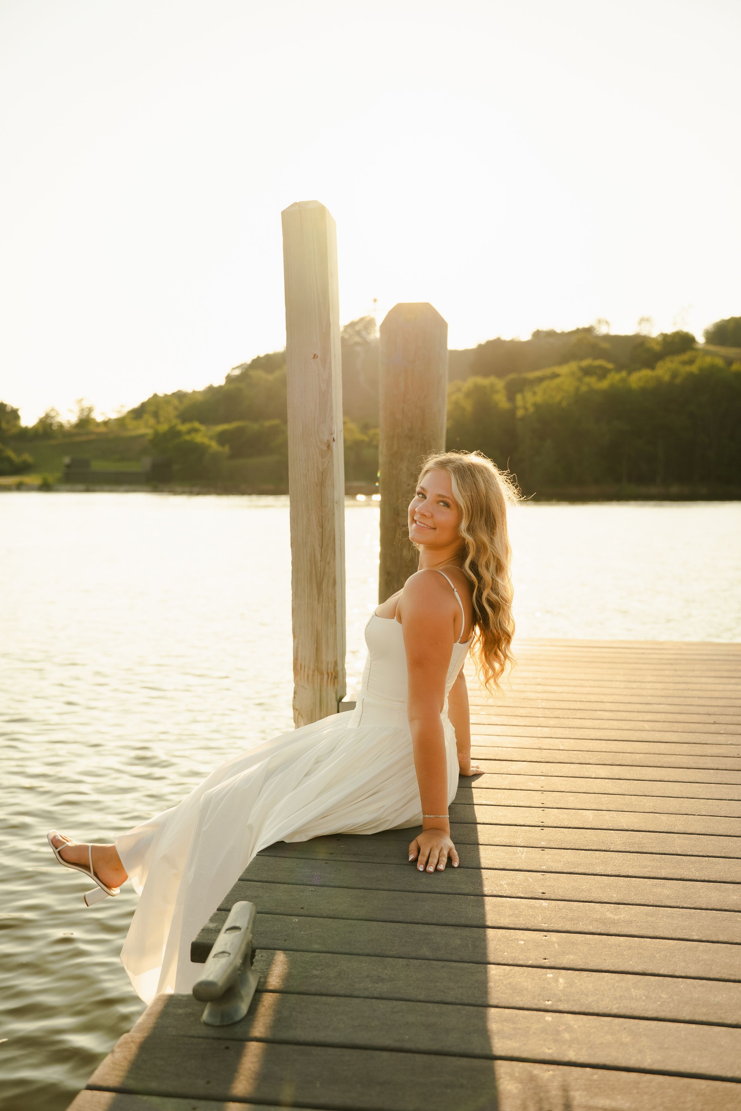 Woman in white dress sitting on a wooden dock by a lake at golden hour sunset.
