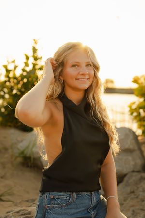 Young blonde woman in black sleeveless top and jeans smiling at sunset on sandy beach with golden backlight.