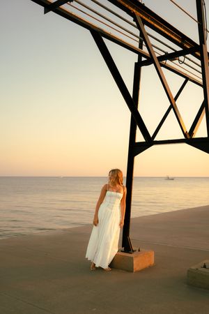 Woman in white dress standing by metal pier structure at golden sunset over calm ocean waters.