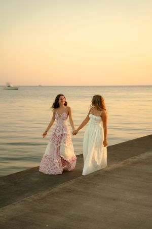 Two women in elegant dresses hold hands on a pier at sunset over calm ocean waters.