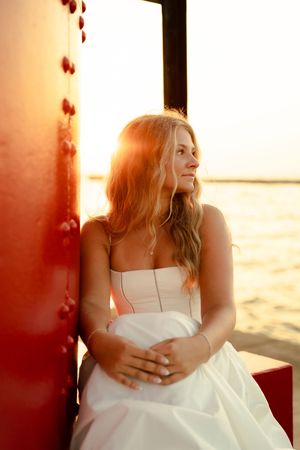 Bride in white dress leaning against red structure at sunset by the water, golden light glowing behind her.