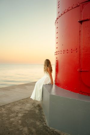 Woman in white dress sits beside a red lighthouse at sunset overlooking a calm ocean.
