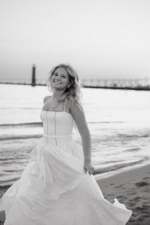 Smiling bride in white wedding gown poses on beach near pier in elegant black and white photo.