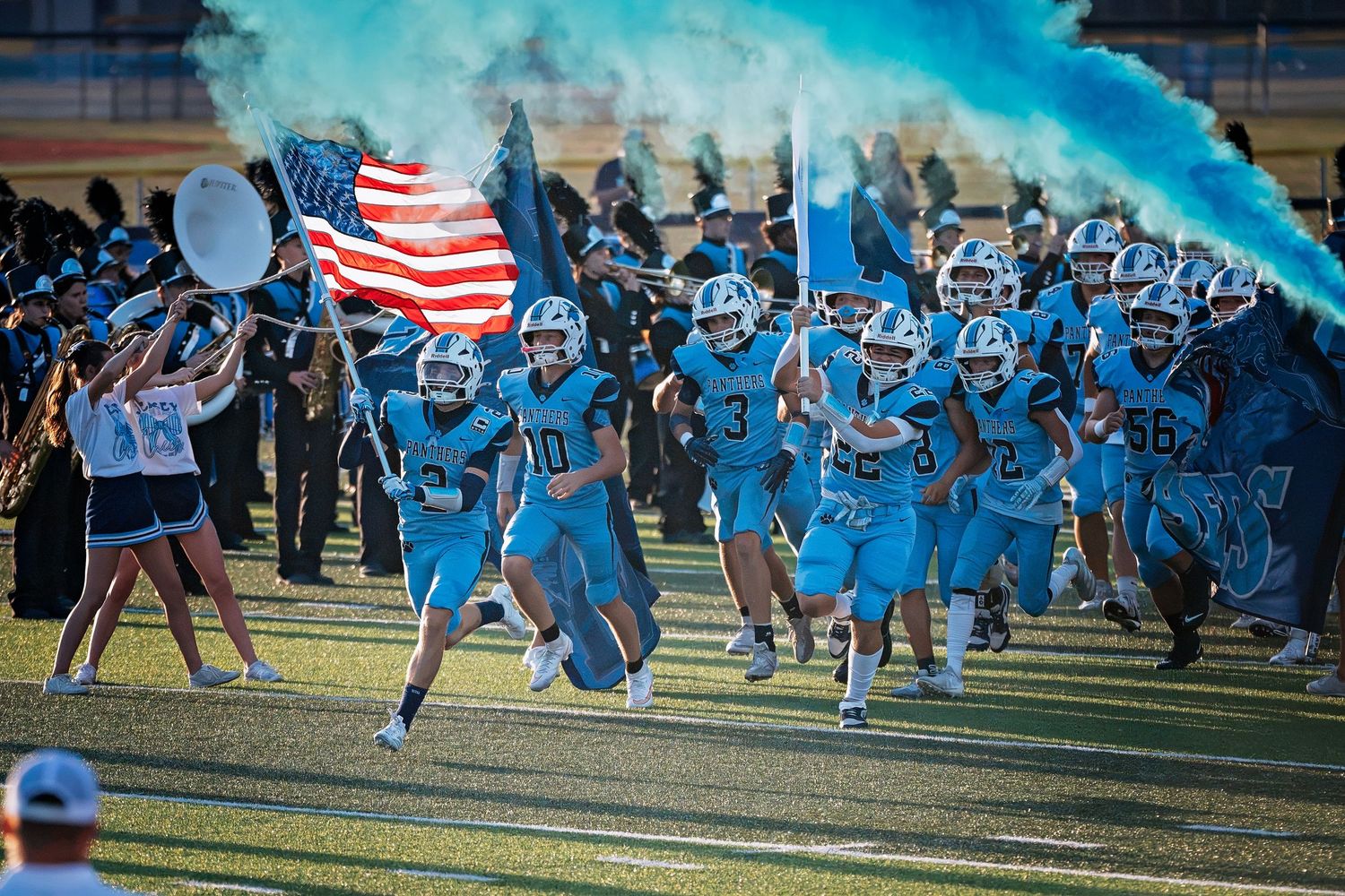 High school football team runs onto field with American flag and teal smoke during pregame entrance ceremony.