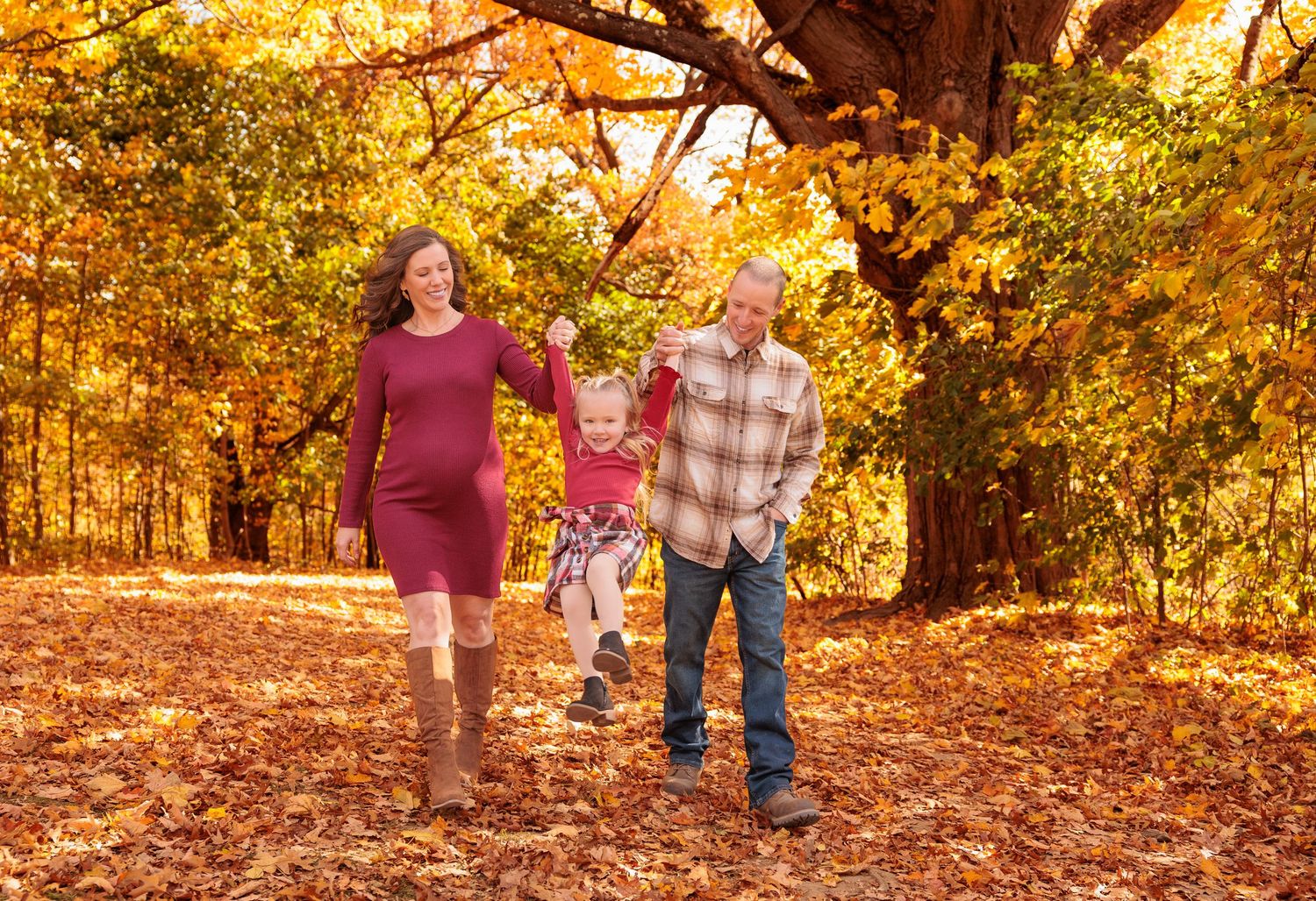 A pregnant mother and father swing their young child between them on a leaf-covered autumn path.