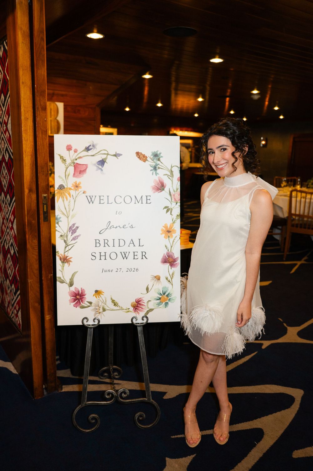 Bride smiling beside floral Welcome to Jane's Bridal Shower sign, wearing white feather-trimmed dress.