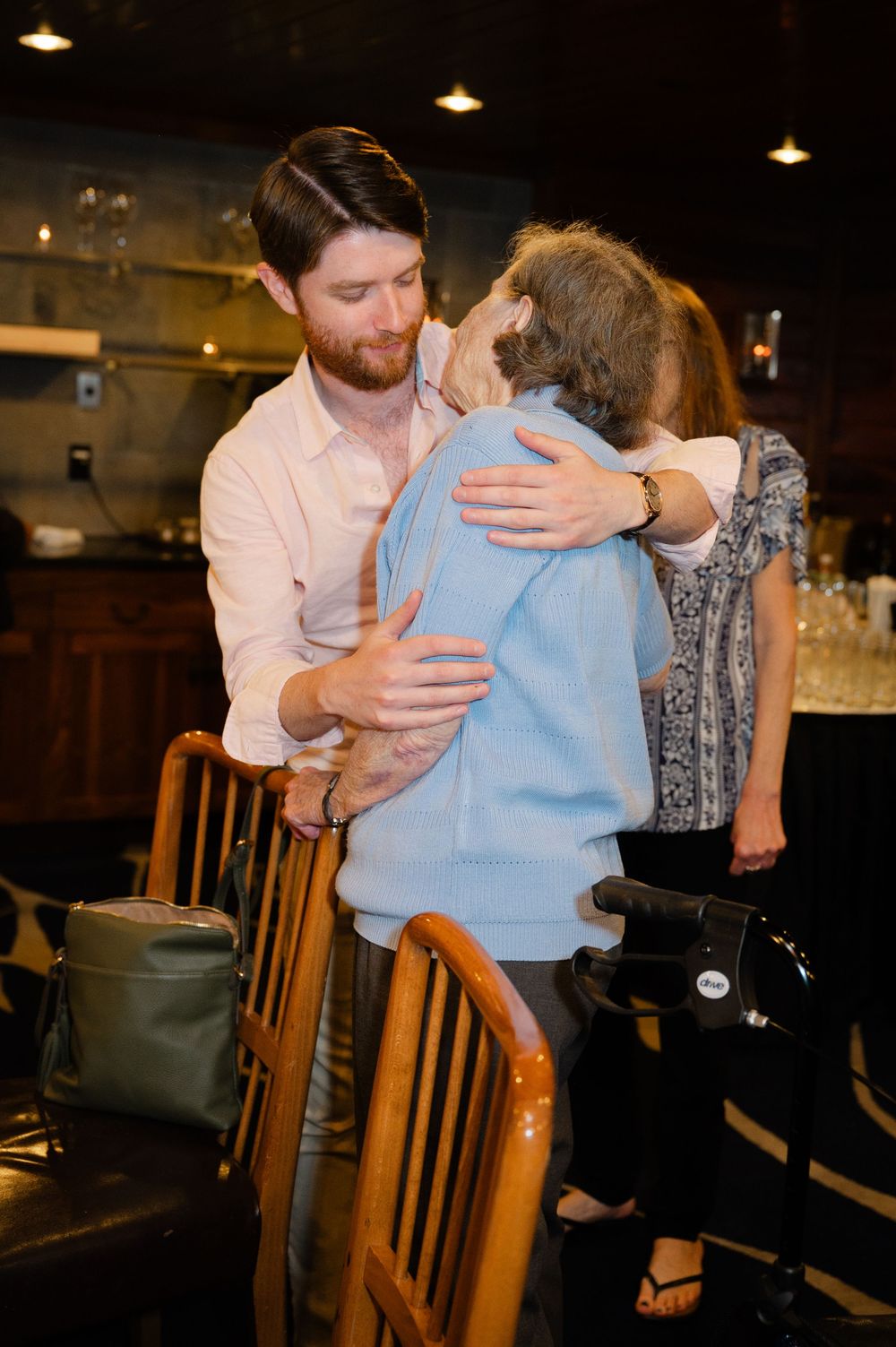 Young man hugging an elderly woman in a restaurant, sharing a warm embrace near wooden chairs.