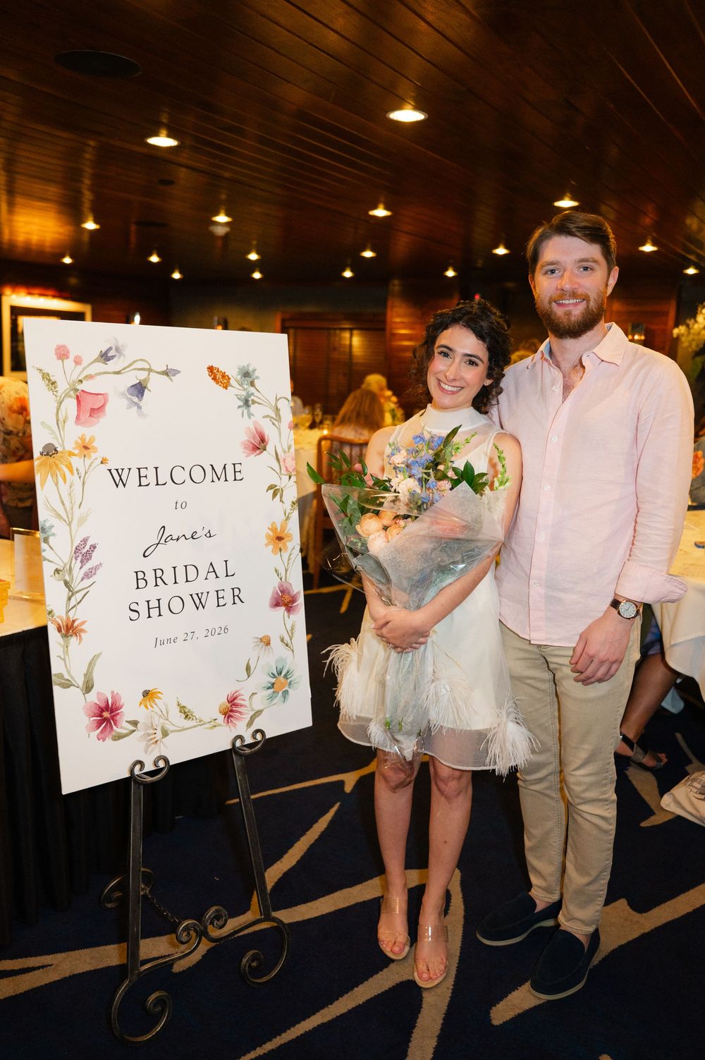 Couple posing together at Jane's Bridal Shower, woman holding floral bouquet beside decorative welcome sign.